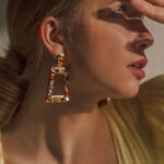 Perfect Outfit - Feminine young lady in trendy dress with low neckline and elegant earrings covering eyes with hand while looking on bright light in studio