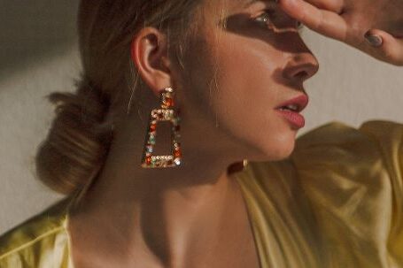 Perfect Outfit - Feminine young lady in trendy dress with low neckline and elegant earrings covering eyes with hand while looking on bright light in studio