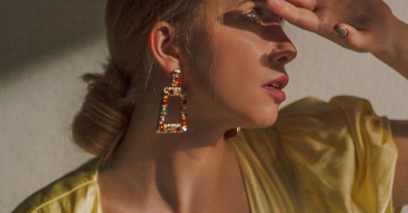 Perfect Outfit - Feminine young lady in trendy dress with low neckline and elegant earrings covering eyes with hand while looking on bright light in studio