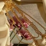 Fashion Inspiration - From above of golden chain with pendant placed on crumpled fabric with mirror near pink flowers in light stylish room