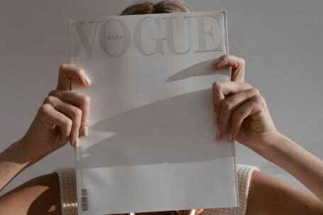 Professional Fashion Tips - Unrecognizable female with big chain on neck standing on white background while covering face with fashion magazine in sunlight