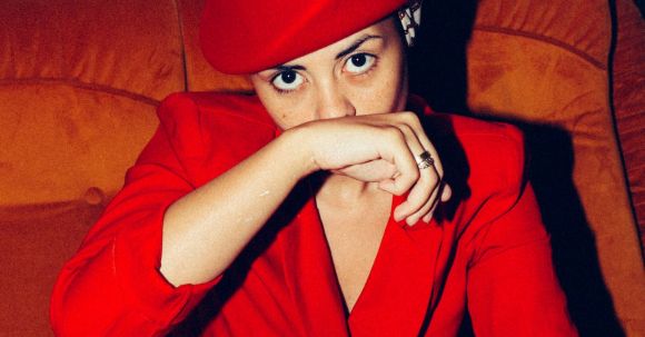 Winter Chic - From above of determined female model wearing trendy suit and retro hat sitting in comfortable armchair and covering mouth with hand while looking at camera