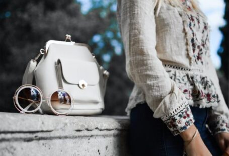 Fashion - woman wearing beige and red floral top leaning on gray concrete slab with white leather bag ontop